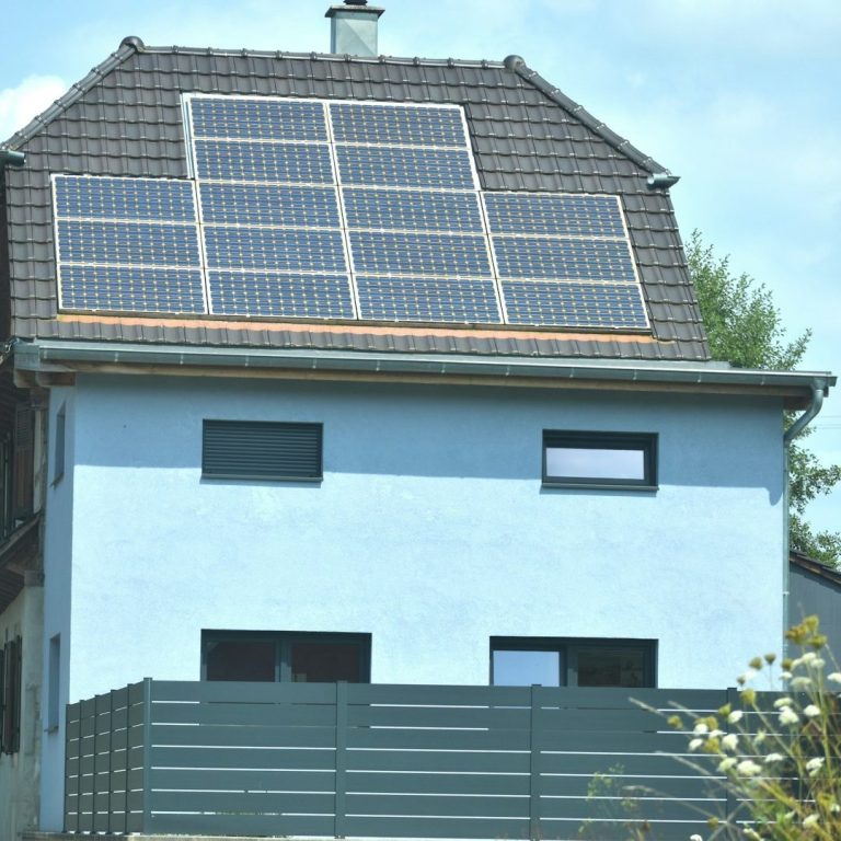 Bild von IONOS Helles Wohnhaus mit Solarpanelen auf dem Dach und Gartenzaun im Vordergrund.